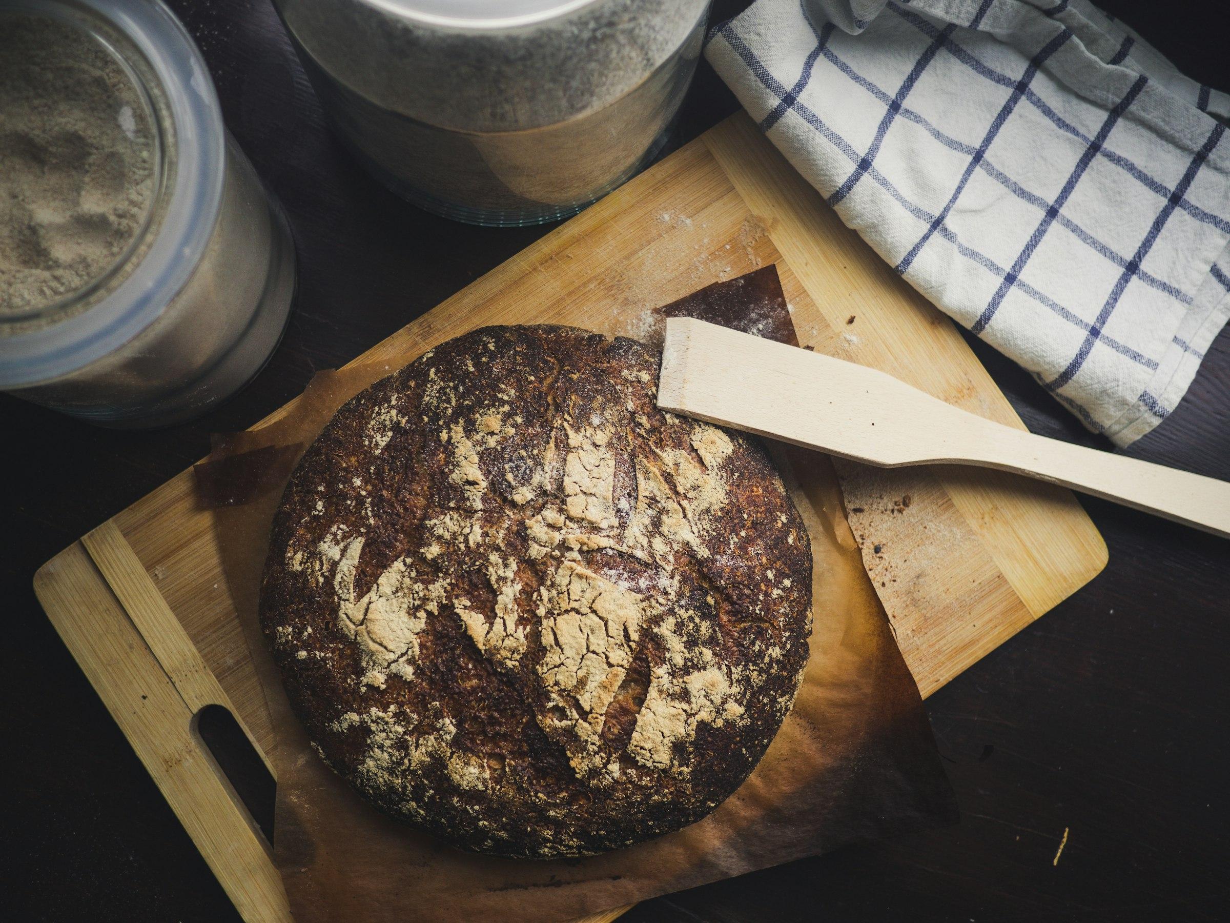 A round loaf of freshly baked bread on a wooden chopping board with a wooden spatula and flour jars nearby