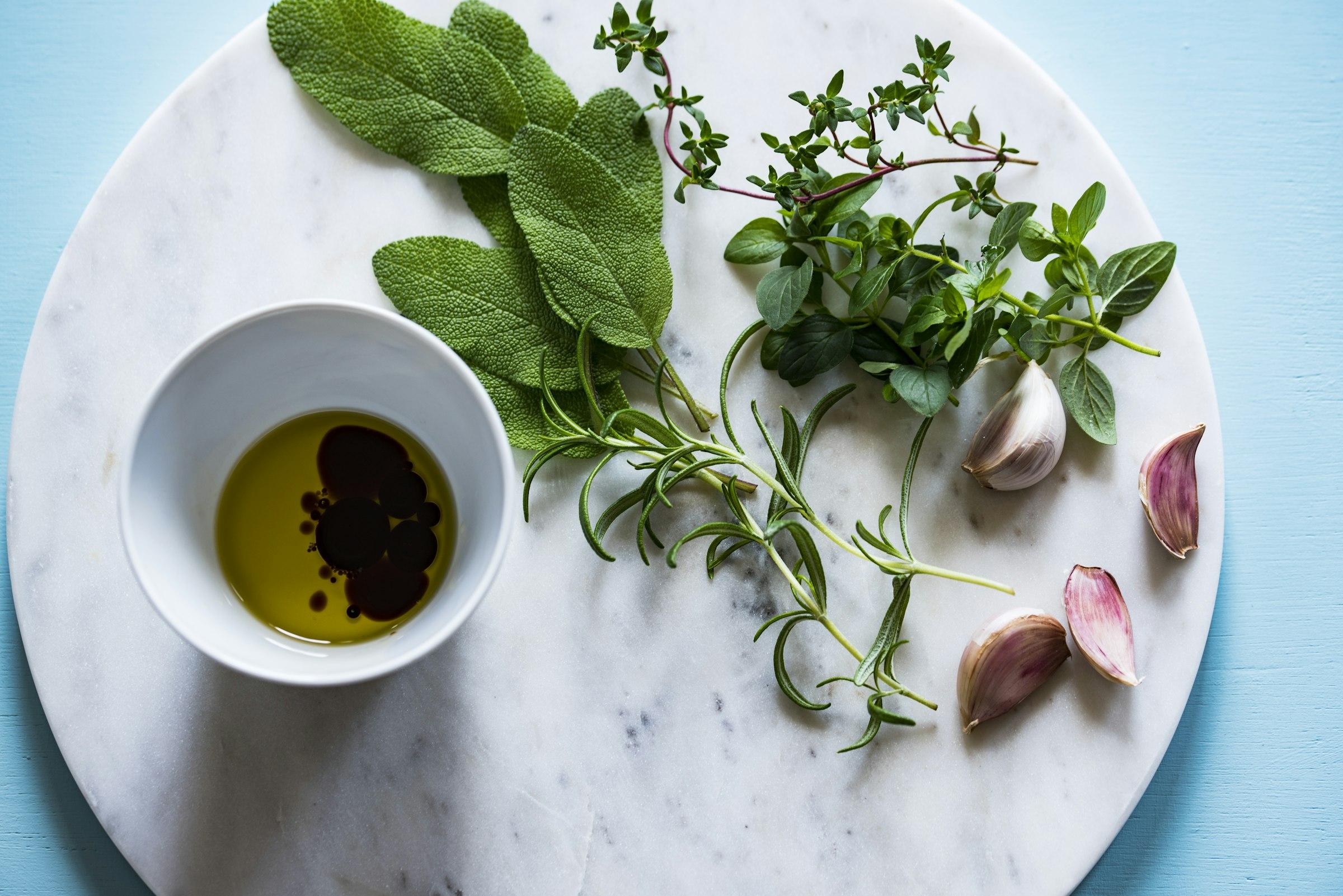 Flat lay of fresh sage, thyme, rosemary, garlic, and olive oil arranged on a marble board